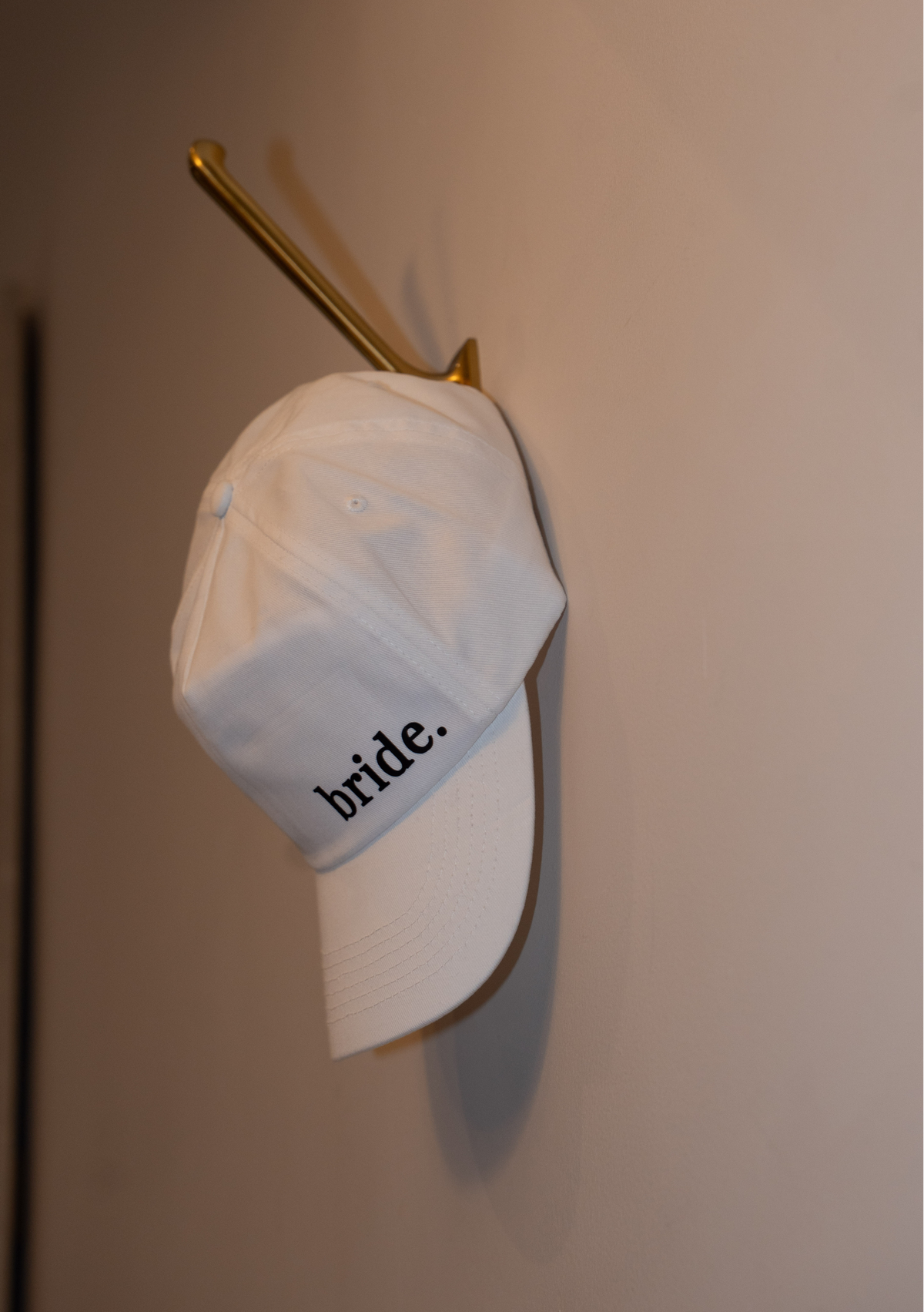 White cap with 'bride' text on a gold hook against a beige wall