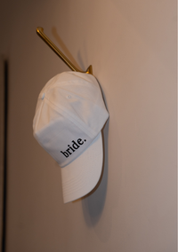 White cap with 'bride' text on a gold hook against a beige wall