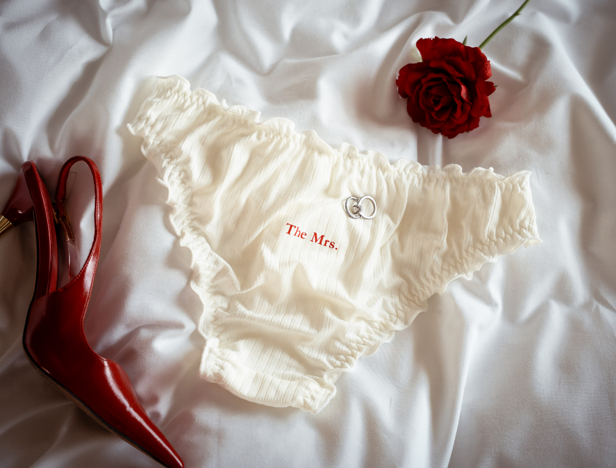 White frill underwear with 'The Mrs.' branding on a white fabric background with a red rose and red stiletto heel.